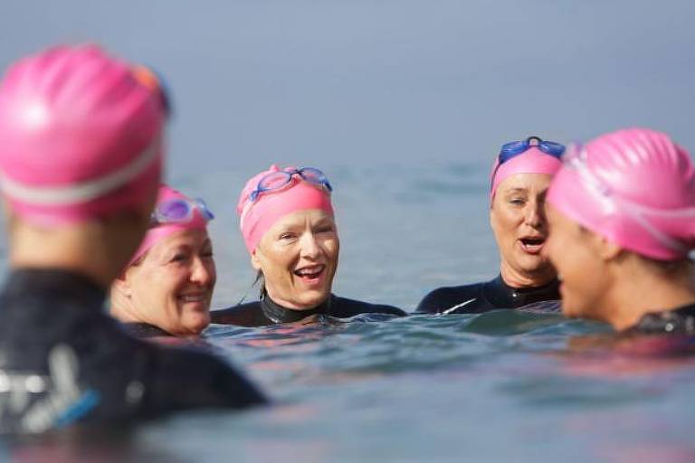 Foto mostra cinco mulheres brancas, com rugas no rosto, nadando ao ar livre. Todas usam tocas rosas e roupas pretas. Elas também estão com óculos de natação na cabeça. As mulheres conversam e riem umas com as outras