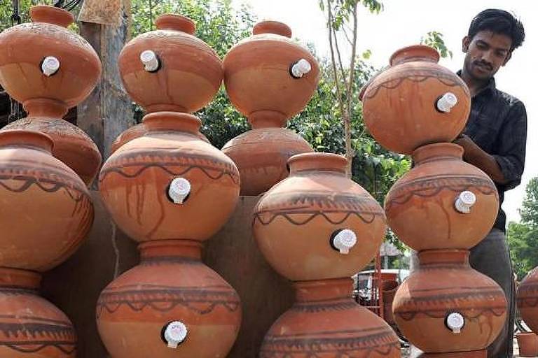 Um homem indiano cuidadosamente arranja uma coleção de potes de barro tradicionais, exibindo a beleza e a simplicidade da cerâmica artesanal. Os potes apresentam desenhos geométricos e linhas onduladas, com tampas arredondadas e torneirinhas brancas.