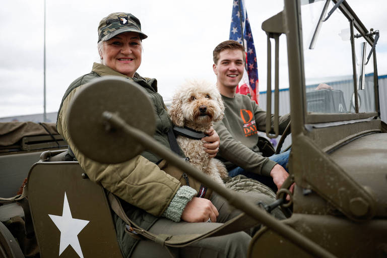 Uma mulher, um rapaz e um cachorro compartilham um momento alegre a bordo de um veículo militar clássico, com a bandeira dos Estados Unidos ao fundo.