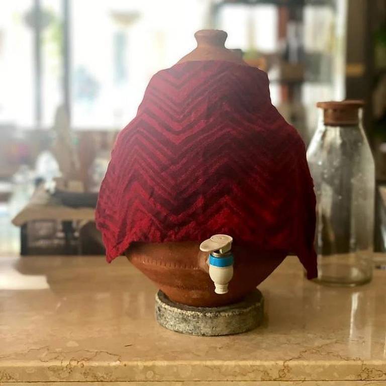 Um pote de barro sobre um balcão. O objeto está coberto por um manto vermelho. Ao fundo, uma garrafa de vidro e um recipiente de madeira.