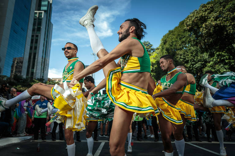 Dançarinos vestidos com trajes nas cores verde e amarelo, inspirados nas animadoras de torcida, dançam na avenida Paulista durante a Parada LGBT; o destaque vai para o dançarino central, que com um sorriso contagiante e uma perna elevada, captura a essência vibrante do evento