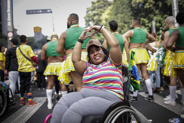 Uma pessoa em uma cadeira de rodas exibe um gesto de coração com as mãos, sorrindo alegremente no meio de uma celebração. Ao fundo, participantes vestidos com trajes amarelos e verdes dançam e celebram