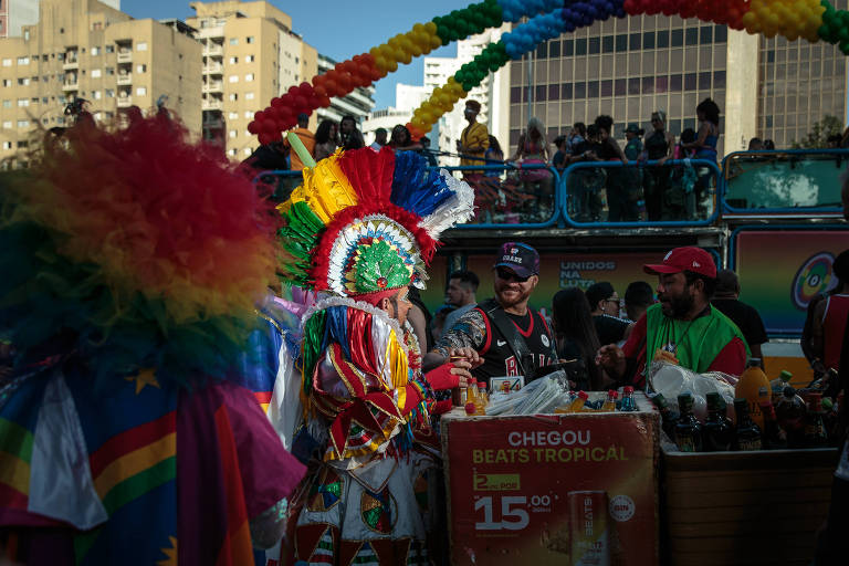 A imagem captura um momento de festividade, onde uma pessoa vestida com um traje colorido e uma máscara elaborada está em primeiro plano, participando de um desfile. Ao fundo, outras pessoas podem ser vistas em um ambiente festivo, com estruturas decorativas e bandeiras coloridas que adornam o cenário urbano.