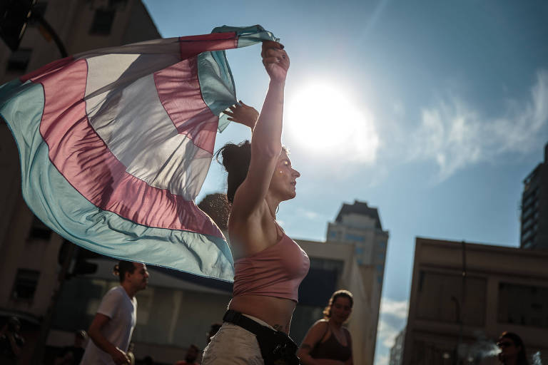 Uma pessoa está segurando uma bandeira trans em um ambiente urbano， com o sol brilhando atrás dela. A bandeira é composta por faixas de cores azul， rosa e branca. A pessoa está vestindo uma blusa de alças e parece estar em movimento， com outras pessoas visíveis ao fundo.