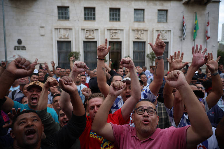 A imagem mostra uma multidão de pessoas levantando os braços e apontando para cima, aparentemente comemorando ou participando de um evento. Ao fundo, há um prédio com várias janelas e bandeiras hasteadas. Algumas pessoas estão usando camisetas de times de futebol.
