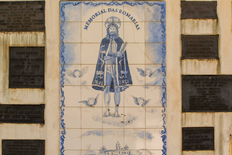 A imagem mostra um painel de azulejos com a inscrição "MEMORIAL DAS ROMARIAS" e a representação de uma figura religiosa, possivelmente um santo, cercado por anjos. Ao redor do painel, há várias placas comemorativas em homenagem a eventos e indivíduos