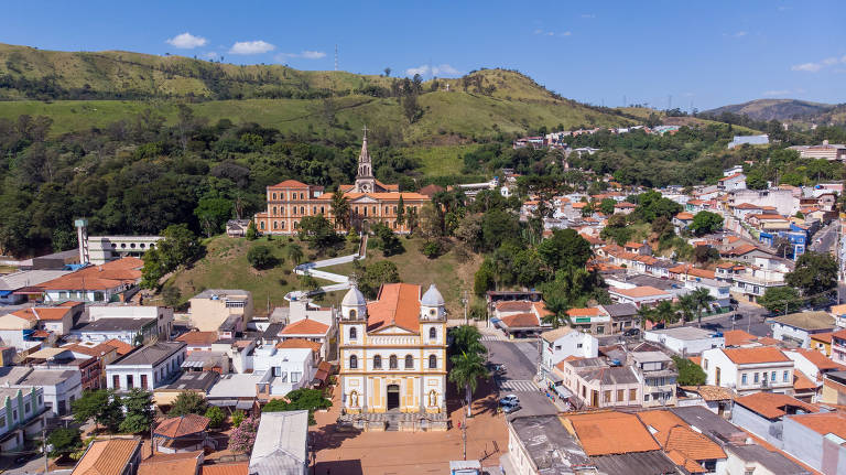 A imagem captura uma cidade histórica, com uma igreja de fachada amarela e detalhes arquitetônicos clássicos em destaque. Ao fundo, colinas emolduram o cenário urbano