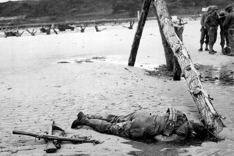 Rifles cruzados na areia ao lado do corpo de um soldado americano na Normandia