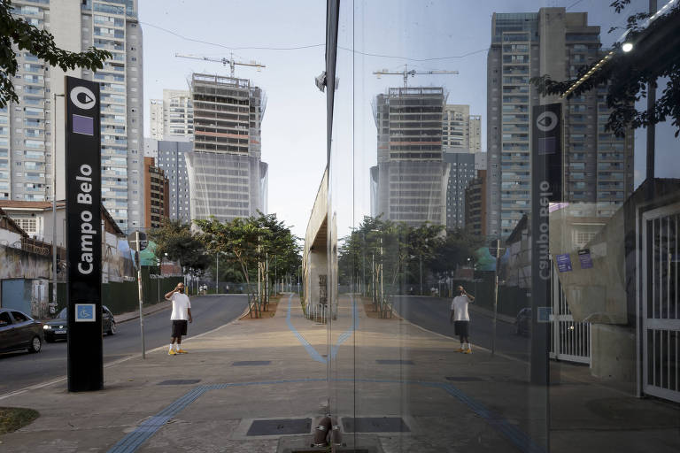 Imagem mostra homem ao lado de totem com o nome da estação Campo Belo e a imagem está refletida no vidro do acesso à estação