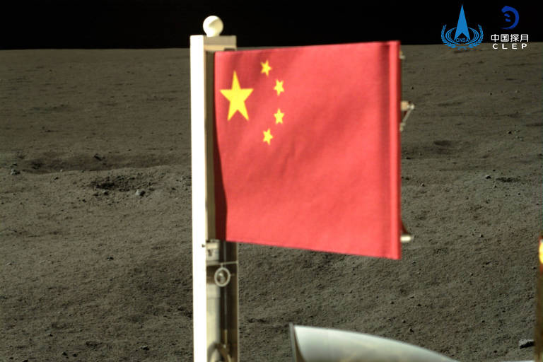 A bandeira vermelha da China com cinco estrelas amarelas é exibida na superfície lunar. O solo cinza e árido da Lua serve como pano de fundo, enquanto parte de uma sonda espacial pode ser vista no canto inferior direito da imagem.
