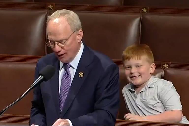 Um homem vestindo um terno e gravata está falando ao microfone em um plenário, enquanto um menino ruivo, com um sorriso contagiante, espreita por trás dele, parecendo compartilhar um momento de diversão.