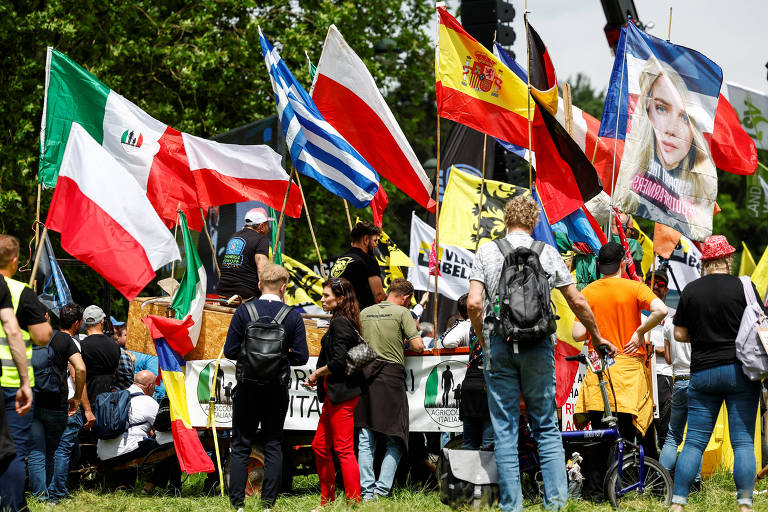 Um grupo diversificado de manifestantes reúne-se ao ar livre, empunhando bandeiras de várias nações e cartazes com mensagens, demonstrando união e diversidade em suas causas.