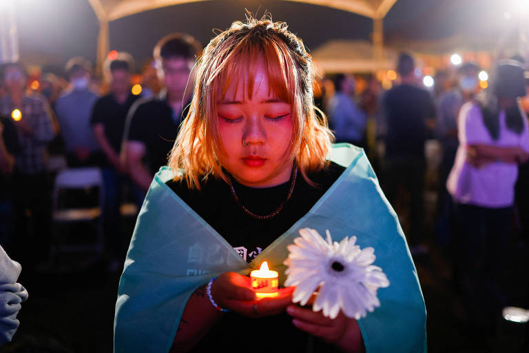 Uma jovem com uma expressão serena e contemplativa segura uma vela acesa e uma flor branca, envolta em uma bandeira, em meio a uma vigília iluminada por velas ao fundo, simbolizando respeito e memória.