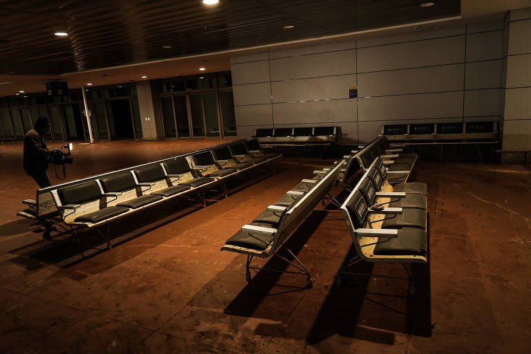 A imagem mostra uma sala de espera  de areporto com fileiras de assentos metálicos vazios. A sala, assim como os assentos, está suja de lama.