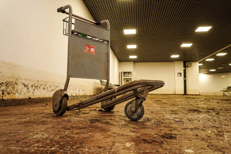 Carrinho de bagagem no saguão sujo pela lama; os bancos de espera estão sujos com detritos como barro, folhas e pequenos galhos, e a bem iluminada sala de desembarque estava às escuras