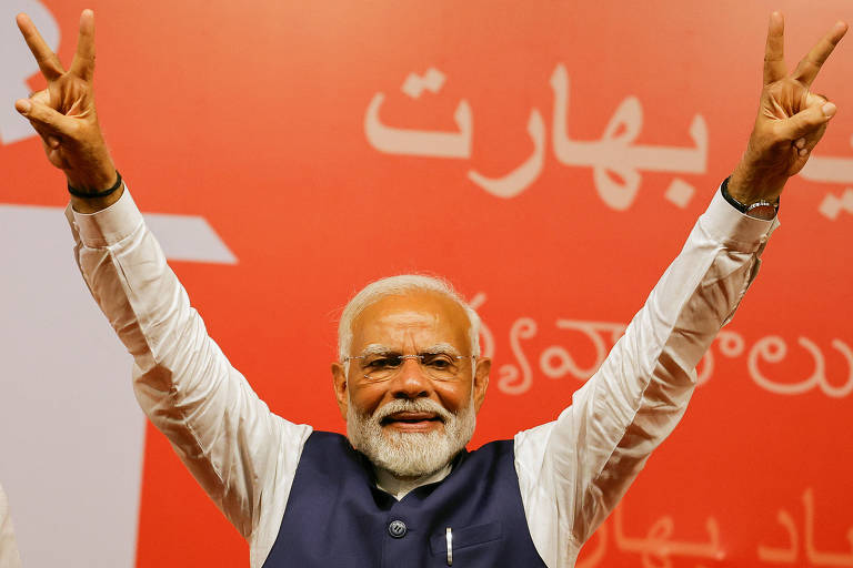 Sorridente, Narendra Modi, premiê da Índia, levanta os braços em um gesto de vitória ou paz, com os dedos em "V", diante de um fundo vermelho com inscrições em hindi. Ele veste um colete sobre uma camisa branca e exibe uma expressão de alegria e confiança.