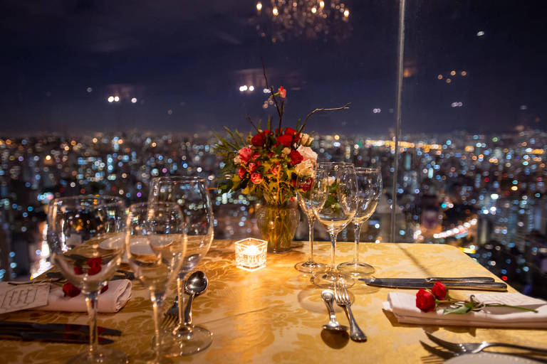 Uma mesa de jantar posta com taças de vinho e um arranjo de flores vermelhas oferece uma vista da cidade de SP iluminada à noite