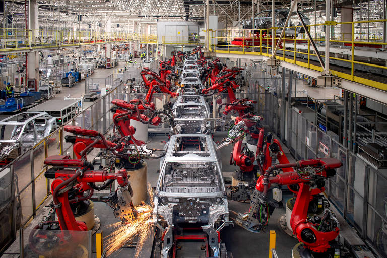 A imagem captura uma linha de montagem de veículos moderna, onde braços robóticos vermelhos estão ativamente engajados na montagem de carros. 