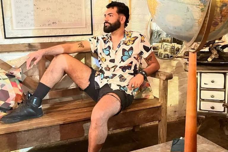 Um homem com barba e cabelo curto, vestindo uma camisa estampada e shorts, está sentado descontraidamente em um banco de madeira. Ele parece estar em um ambiente com decoração vintage, incluindo um globo terrestre ao seu lado