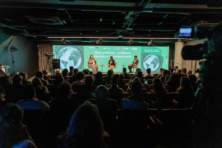 Três pessoas em cima de um palco de um auditório falam para uma platéia cheia. Atrás delas, um telão luminoso na cor verde, com imagens do planeta Terra