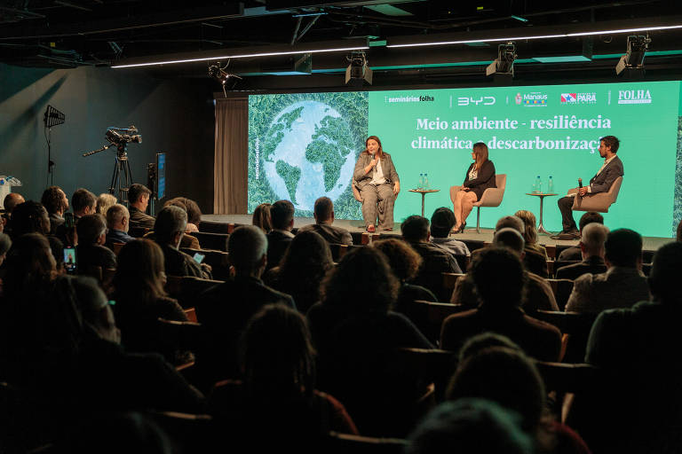 Duas palestrantes e um mediadorcompartilham suas ideias em um palco iluminado, diante do público do evento. O painel atrás do palco é verde e tem um mapa-múndi ao fundo.