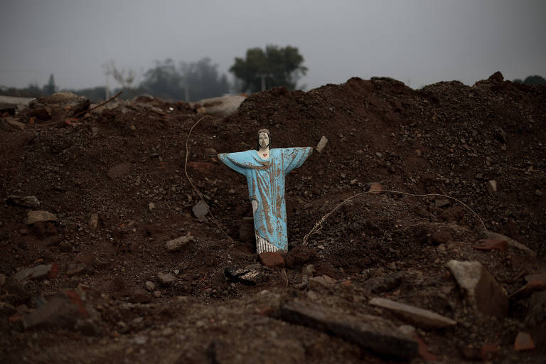 Uma escultura em formato de cruz, adornada com um tecido azul, se ergue em meio a um terreno devastado, com montes de terra e destroços ao fundo sob um céu nublado