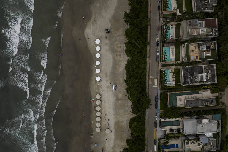 Fotografia feita de cima mostra, do lado esquerdo, o mar e as ondas e, do lado direito, prédios à beira mar. Na faixa de areia é possível ver guarda-sóis brancos espalhados pela praia.
