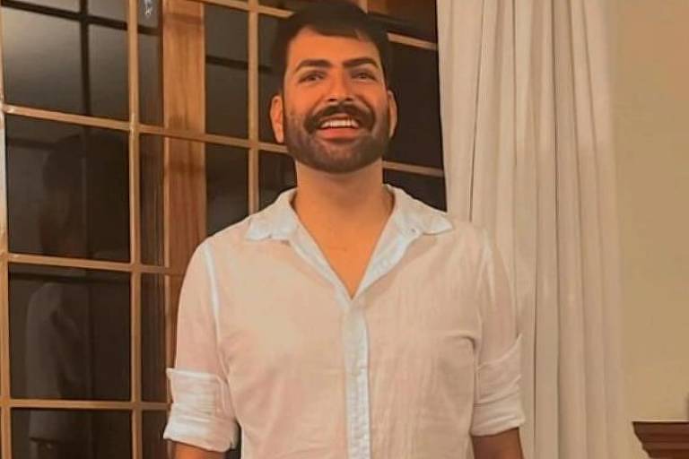 Um homem com barba e cabelo curto posa para a foto com um sorriso leve, vestindo uma camisa branca de mangas curtas e calças brancas, em um ambiente interno com decoração clássica e uma janela ao fundo
