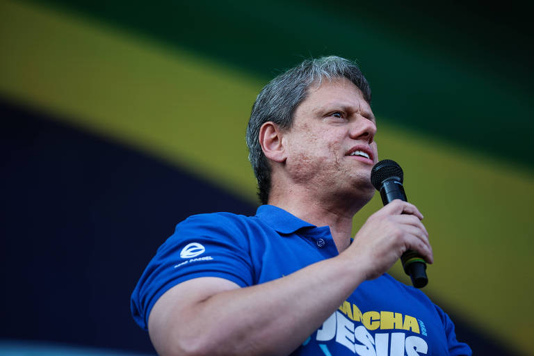 Imagem de um homem segurando um microfone e falando. Ele está vestindo uma camiseta azul com as palavras 'Marcha para Jesus' visíveis. Ao fundo, há uma faixa com as cores verde e amarela.