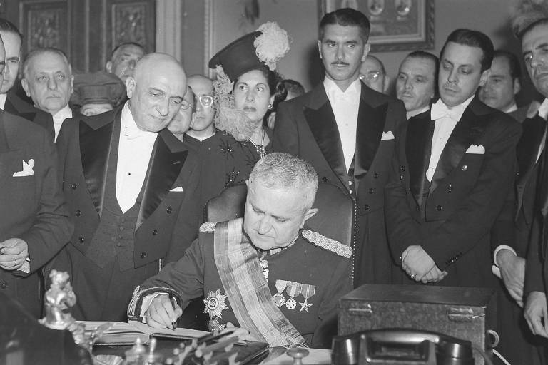 Eurico Dutra, em foto de 1946, cercado por assessores e outros homens, ele assina documento e usa uniforme militar com medalhas e uma faixa presidencial que cruza o peito