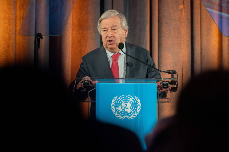 Um homem vestindo terno e gravata fala em um púlpito com o emblema das Nações Unidas, diante de uma audiência atenta. Duas cabeças estão desfocadas em primeiro plano da imagem.