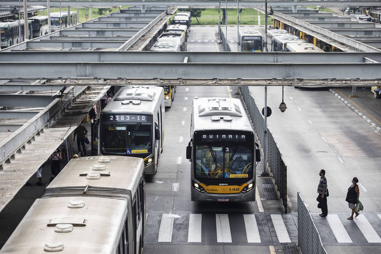 Um ônibus com pintura amarela, no centro da imagem, é visto em frente a uma faixa de pedestres e ao lado de uma fileira de outros ônibus estacionados numa faixa de asfalto; uma estrutura metálica com vigas e marquises aparece na imagem sobre os veiculos