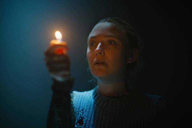 Uma jovem com expressão atenta segura uma vela acesa, iluminando seu rosto em um ambiente escuro.