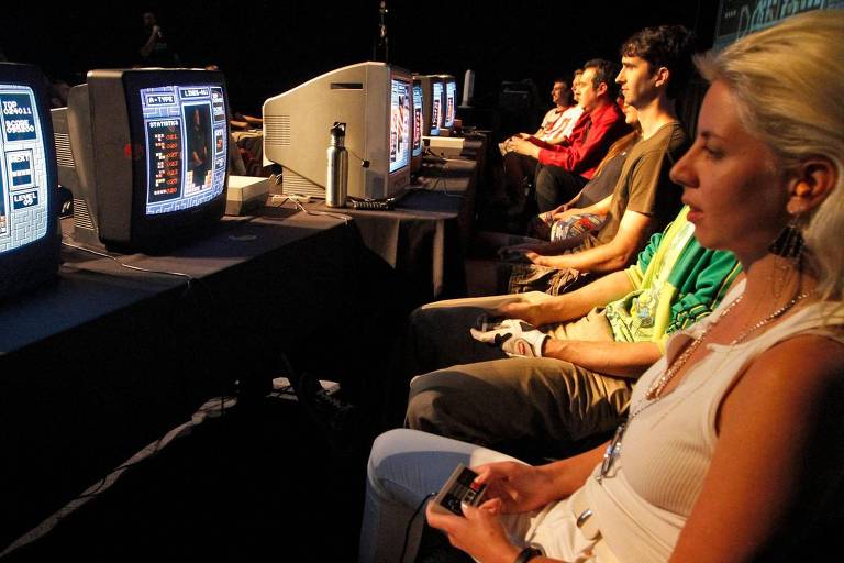 Um grupo de pessoas está sentado em uma fileira, concentrado em jogos de vídeo game clássicos em monitores antigos. A iluminação ambiente é baixa, destacando as telas.