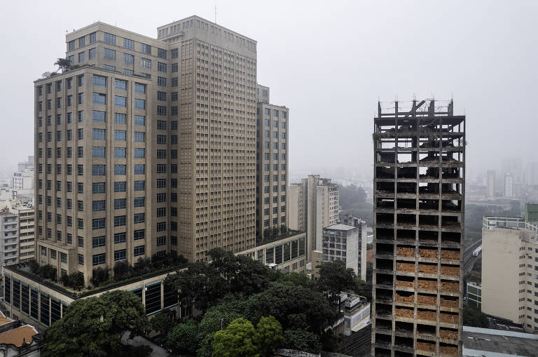 A imagem captura um edifício inacabado e abandonado, com estruturas de concreto expostas e paredes de tijolos marcadas por pichações. Ao fundo, a paisagem urbana densa e nebulosa de uma cidade, com prédios residenciais e comerciais, sugere uma metrópole em constante expansão. Uma via férrea serpenteia ao lado do edifício, adicionando um elemento dinâmico à cena urbana.