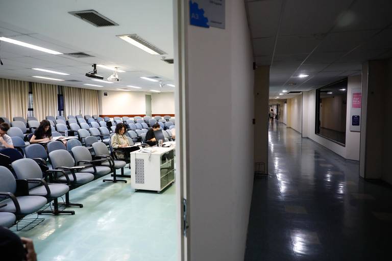 A imagem mostra uma sala de espera à esquerda, com várias cadeiras azuis ocupadas por pessoas. À direita, há um corredor vazio com piso brilhante e paredes claras. O ambiente parece ser de um edifício público ou hospitalar.
