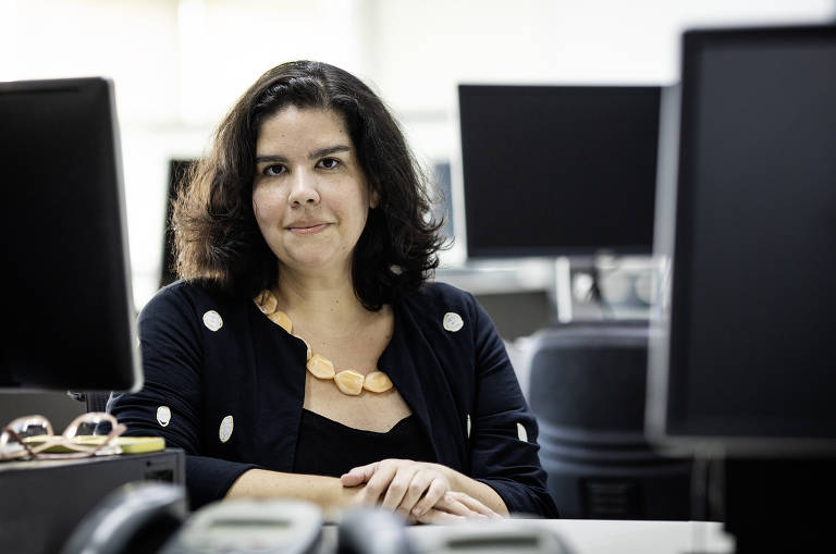 Mulher está sentada em sua mesa em um escritório iluminado, cercada por monitores de computador. Ela veste um casaco escuro com botões claros e um colar de tons amarelos.