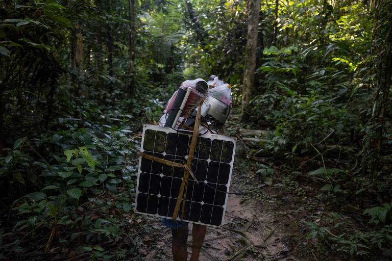 Homem caminha pela floresta tropical, carregando painel e satélite da Starlink amarrado às costas com cordas. 