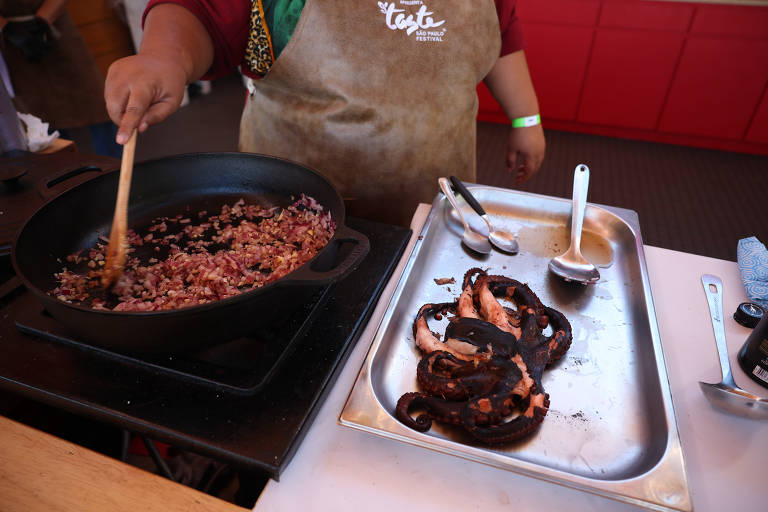Uma cozinheira em um avental está preparando carne em uma frigideira sobre o fogão, com ingredientes e utensílios de cozinha ao redor, incluindo pimentões pretos em uma bandeja branca ao lado.