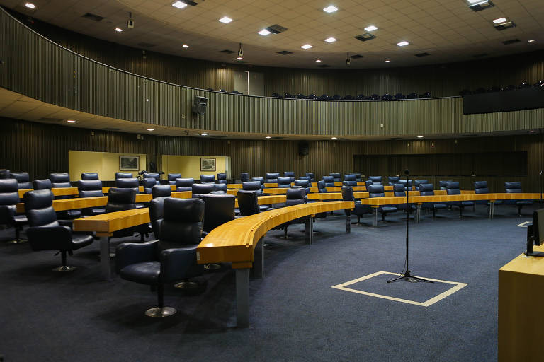 A imagem mostra a Câmara Municipal de São Paulo, um anfiteatro moderno e vazio, com cadeiras de couro azul escuro e mesas de madeira clara. No centro, há um púlpito com um microfone. As paredes são revestidas com painéis acústicos e há partes elevadas com assentos adicionais