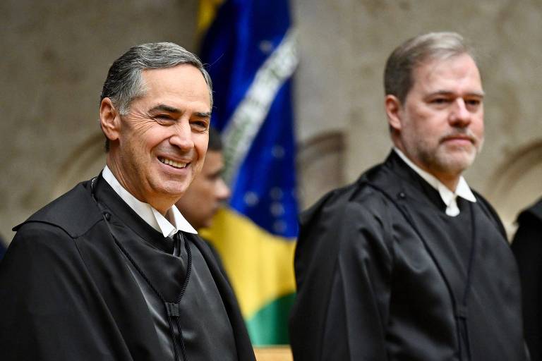 Dois homens brancos de cabelos grisalhos vestidos de togas pretas. Os dois estão de pé e um deles, Barroso, sorri. Ao fundo, uma bandeira do Brasil