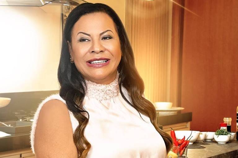 Uma mulher sorridente exibe confiança e elegância em uma cozinha moderna. Vestindo uma blusa de seda sem mangas e calça jeans, ela se apoia em uma bancada de mármore, cercada por utensílios de cozinha e ingredientes, sugerindo uma paixão pela culinária em um ambiente doméstico sofisticado.