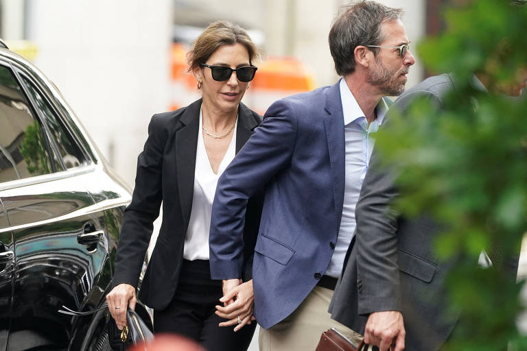 Dois indivíduos, uma mulher e um homem, estão engajados em uma conversa casual na calçada. A mulher veste um blazer preto e óculos escuros, segurando uma bolsa, enquanto o homem está em um blazer azul, com as mãos nos bolsos de sua calça. Eles parecem estar em um momento descontraído, talvez após um evento ou reunião, com um veículo de luxo preto e um motorista ao fundo, indicando um ambiente urbano e sofisticado.