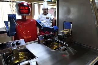 A cook stands behind a robot as it process food at the kitchen of a restaurant in Hefei