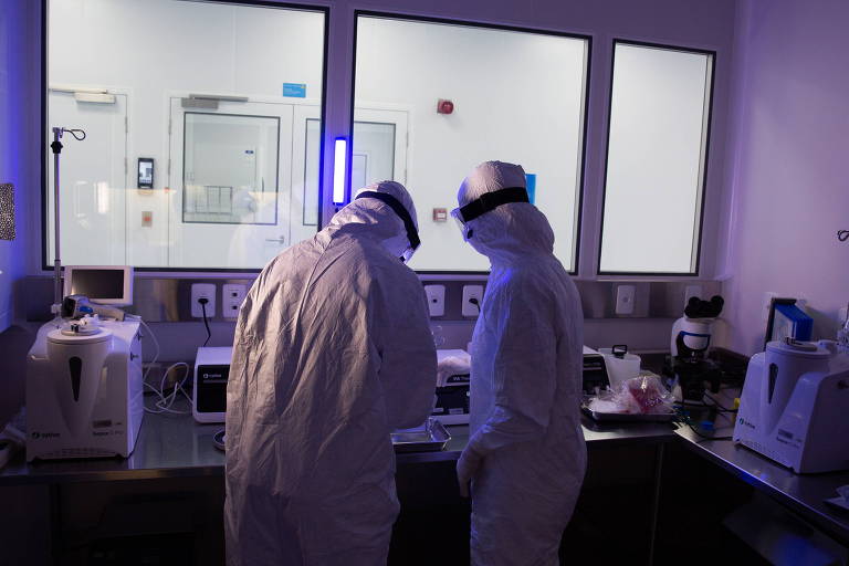 Duas pessoas om roupas de segurança, como jaleco, óculos e touca, estão dentro de um laboratório pouco iluminado
