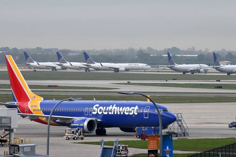 Imagem mostra avião nas cores azul, amarelo e vermelho com o nome da Southwest escrito. Ele está parado em um aeroporto. Ao fundo, é possível ver outros aviões estacionados.