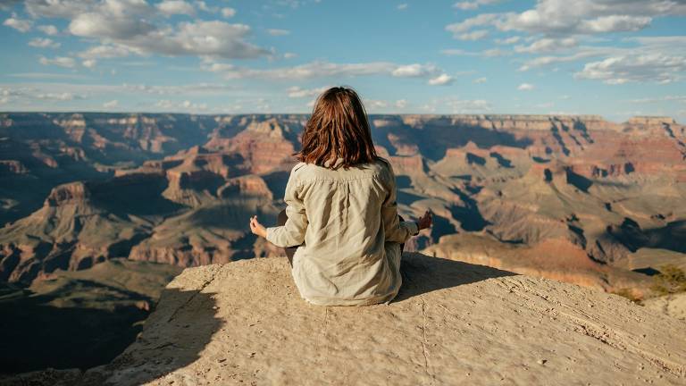 Uma pessoa sentada de costas para a câmera medita em posição de lótus no topo de um penhasco, com uma vista deslumbrante do Grand Canyon ao fundo. A vastidão do cânion e o céu azul com nuvens esparsas criam um cenário de tranquilidade e imensidão, evocando uma sensação de paz e conexão com a natureza.
