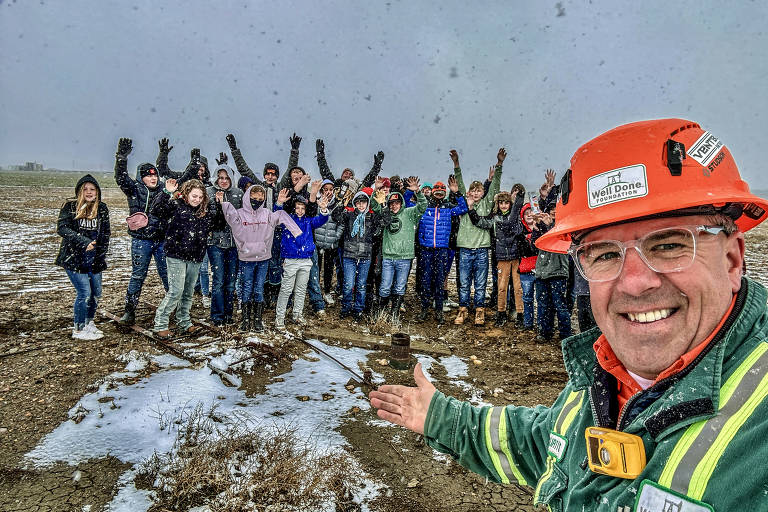 Um homem sorridente com capacete de segurança laranja e óculos tira uma selfie com um grupo animado de pessoas ao fundo. Eles estão em um campo aberto com neve esparsa no chão, levantando os braços em celebração