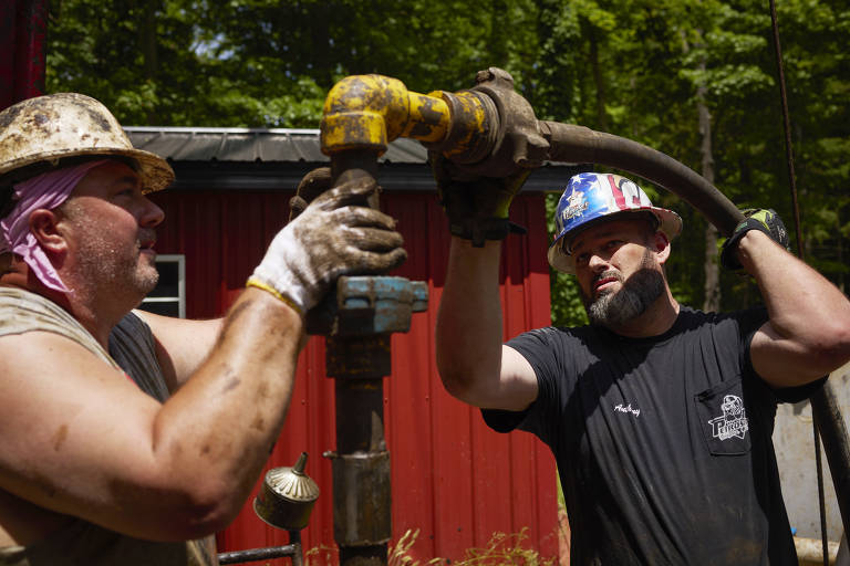 Dois trabalhadores, sujos de graxa e suados, colaboram na operação de uma ferramenta para injetar concreto em poço de petróleo
