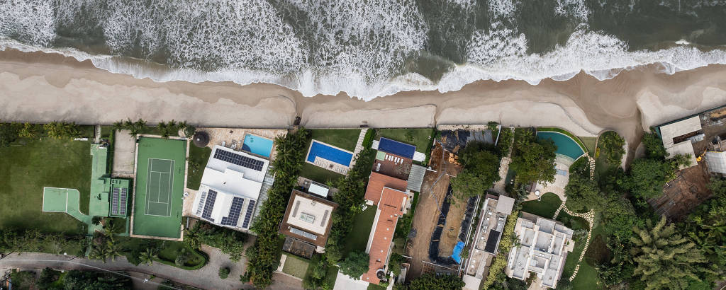 Imagem aérea mostra faixa de areaia banhada pelo mar com casarões na encosta, alguns com piscinas e quadras esportivas